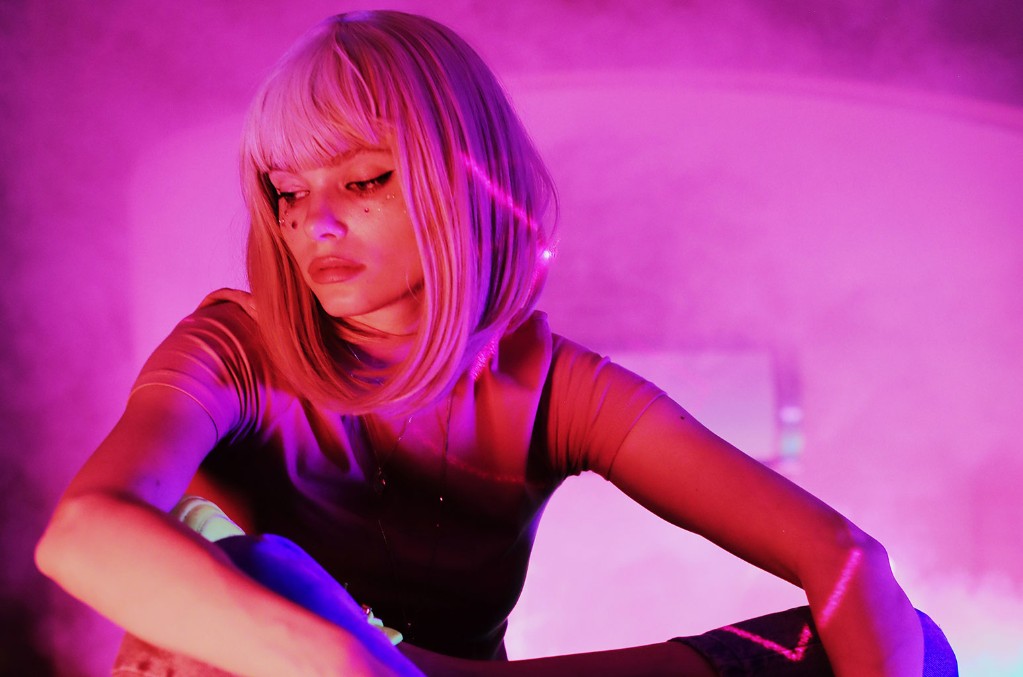 Photo of Winona Oak in a pink wig looking away from the camera against a background lit with pink lights